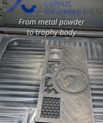 3D printed AU Materials trophy body inside a laser powder bed fusion machine, with text reading “From metal powder to trophy body.